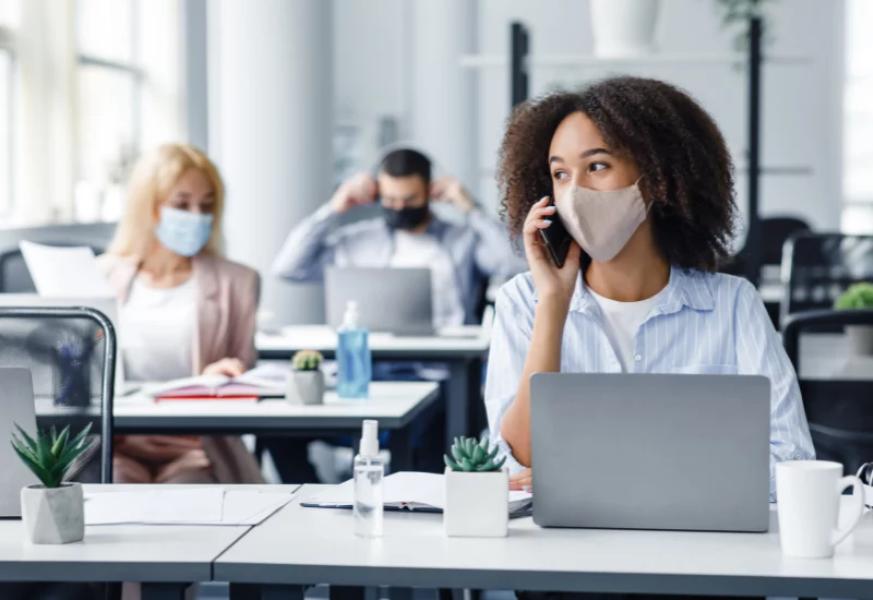 People sat at their desks in the office with masks on and on the phone while having their laptops open.