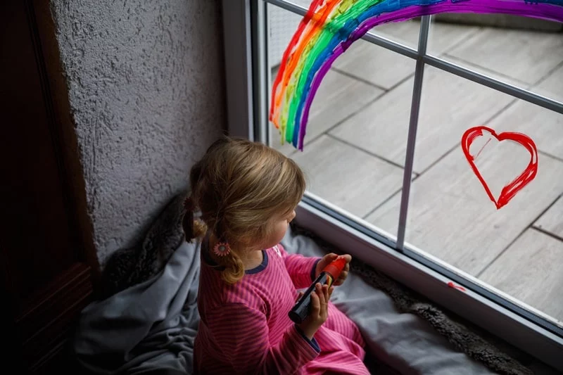 little girl playing with pens and rainbow and heart drawn on glass window
