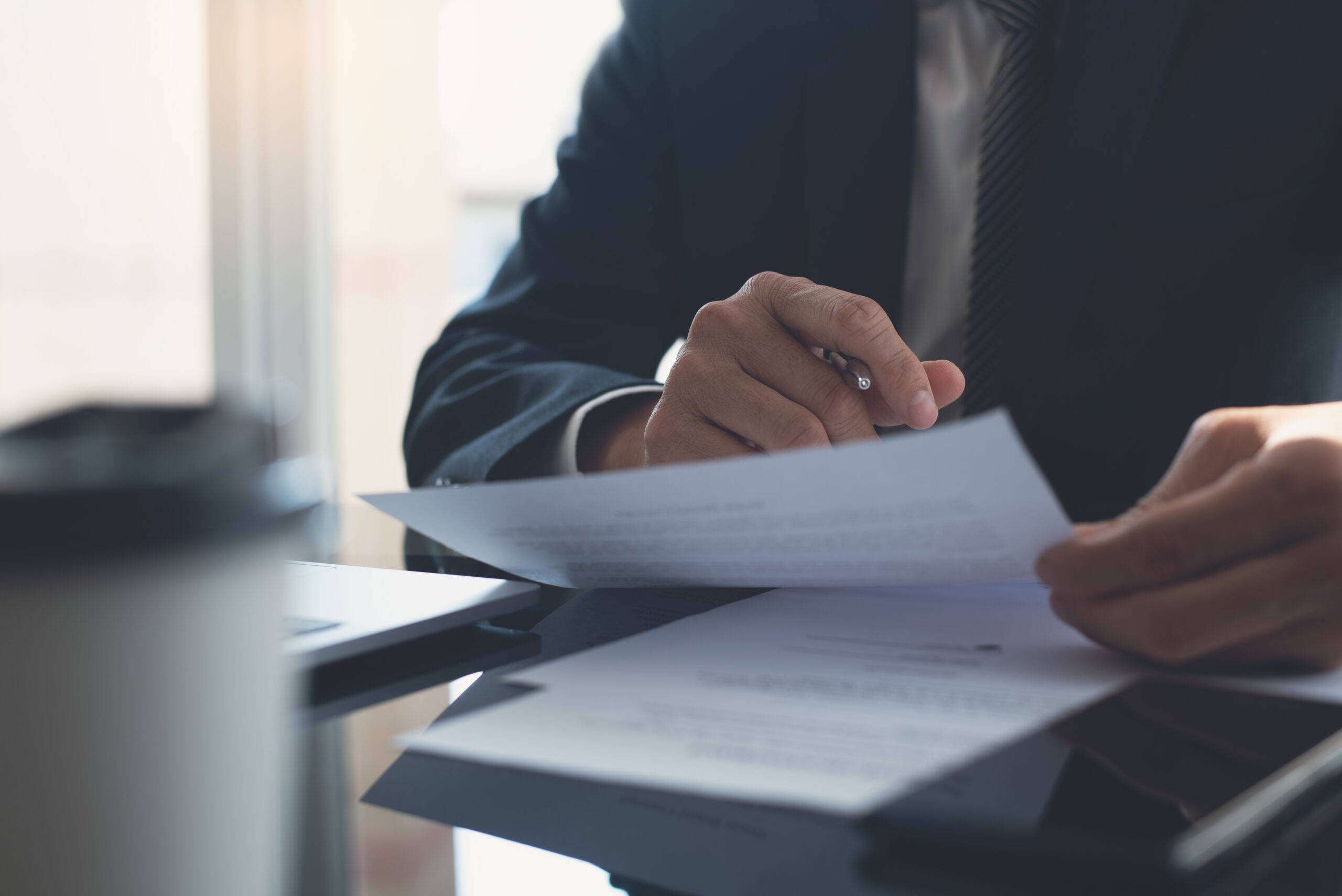Signing contract, business agreement and deal concept. Closeup businessman in black suit reading before signing business contract documents at desk with laptop computer, busy with paperwork. Man manager proofing report at office