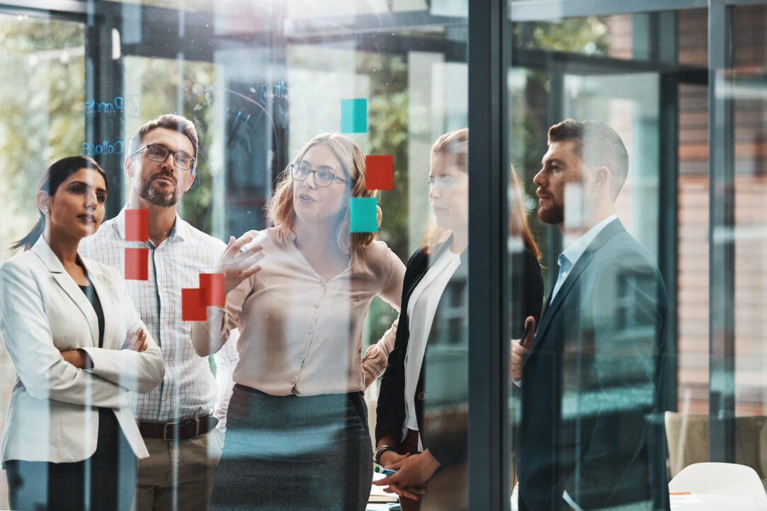 Woman, group and discussion with notes on board with review, listening and planning with brainstorming. Coach, training and team by glass wall, questions and collaboration with strategy in office
